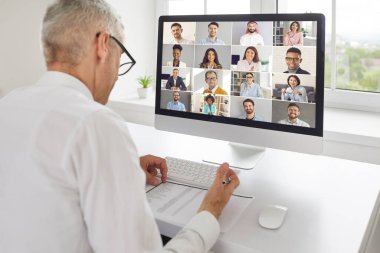 Üst düzey bir işadamı bilgisayar ekranının önünde oturmuş sanal online personel toplantısı yapıyor. Kafkasyalı, siyahi ve Orta Doğulu çeşitli uluslararası çalışma arkadaşlarını dinliyor.