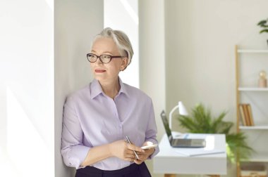 Başarılı bir mastür kadın. Elinde cep telefonuyla gülümseyen kıdemli iş kadını pencereden içeri bakıyor. Ofisinde düşünceli bir şekilde poz veren şık gri saçlı bir kadının portresi..