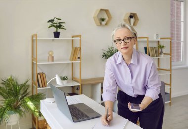 İş görüşmesine hazırlanırken ya da çalışırken not defterine not alan kıdemli iş kadını. Ofiste kendine güvenen olgun bir kadın cep telefonu ve dizüstü bilgisayarını iş için kullanıyor. İş konsepti.