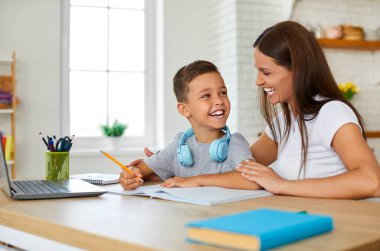 Evdeki neşeli genç anne, birinci sınıftaki oğluna ev ödevini yapması için neşeyle yardım ediyor. Kadın ve küçük çocuk ellerinde dizüstü bilgisayarlarıyla masada oturmuş birbirlerine bakıp gülüyorlar..