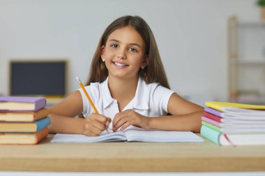 Küçük pozitif kız ilkokul öğrencisi ders kitapları ve not defterleriyle masada poz veriyor ve elinde kalemle kameraya bakıyor, sınıfta oturuyor. Okula dönmek, ilköğretim