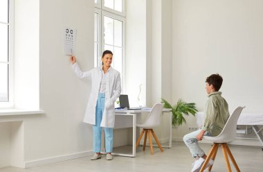 Çocuk klinikte göz muayenesi yaptırıyor. Doktor göz muayenesi yapıyor. Oftalmolog oğlanların görme yeteneğini test ediyor. Modern beyaz tıp bürosunda oturan mutlu okul çocuğu ve göz doktorunun gösterdiği göz çizelgesine bakıyor.