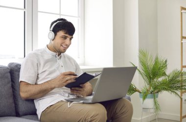 Çevrimiçi öğrenim gören mutlu genç adam kendi evinde yeni yabancı dil öğreniyor. Kulaklıklı bir erkek öğrenci kanepeye oturmuş, modern dizüstü bilgisayara bakıyor, notlar alıyor ve gülümsüyor.
