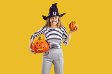 Cadılar Bayramı balkabaklı komik, kızgın cadı kızın stüdyo portresi. Sarı arka planda yalnız duran siyah şapkalı genç bir kadın, iki oyulmuş balkabağı tutuyor ve komik, çılgın bir surat ifadesi sergiliyor.