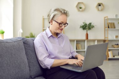 Şık, orta yaşlı, gözlüklü bir kadın rahat ofisindeki kanepede dizüstü bilgisayar yazarak çalışıyor. Açık mor tişört ve pantolon giyiyor. Yaşlı kadın ekrana bakıp gülümsüyor..