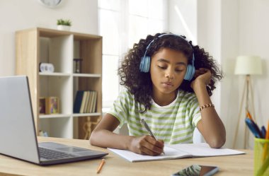 Evde okuyan ciddi bir okul çocuğu. Kulaklıklı Afrikalı bir öğrenci dizüstü bilgisayarla masada oturuyor, deftere yazıyor, ders dinliyor, ödev yapıyor ya da sınava hazırlanıyor.