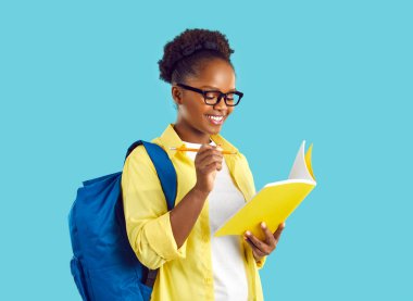 Sarı tişörtlü, gözlüklü, mavi arka planda duran güzel Afro-Amerikan öğrenci kız portresi, deneme metnini not defterine ya da yazıya hazırlamak için sınav planlayıcısında düzenliyor.