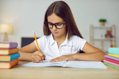 Zeki okul çocuğunun portresi. Gözlüklü küçük kız masada oturuyor, yeni şeyler okuyor ve deftere yazıyor. Öğrenim ve eğitim kavramı