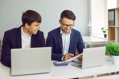 Ofiste çalışan insanlar. İki mutlu genç adam bir masada oturuyor, birlikte evrak işleriyle uğraşıyorlar, bazı finansal belgelere bakıyorlar, modern dizüstü bilgisayarları kullanıyorlar ve konuşuyorlar.