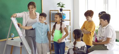 Neşeli arkadaş canlısı çocuklar ve kızlar, ilkokul sınıfında bulunan tahtaya yakın cevap veren akranlarına gülümseyen kadın öğretmenle birlikte tablonun yanında görev yapıyorlar. Panoramik çekim