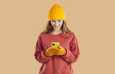 Güz kıyafetleri içinde cep telefonu kullanan güzel bir kadın. Stüdyo portresi. Sıcak kazaklı, şapkalı ve eldivenli güzel sarışın kız. Bej arka planda duruyor, cep telefonuna bakıp gülümsüyor.