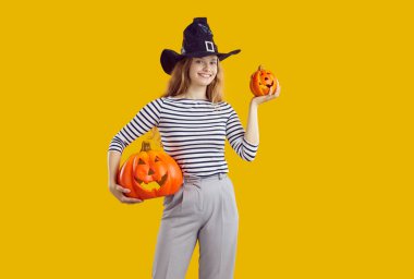 Şapkalı genç ve çekici bir öğrenci elinde iki Cadılar Bayramı balkabağı tutuyor. Jacks fenerlerinin gece parti için hazırlandığını gösteriyor. Cadılar Bayramı tatili, turuncu stüdyo arka planında duruyor.