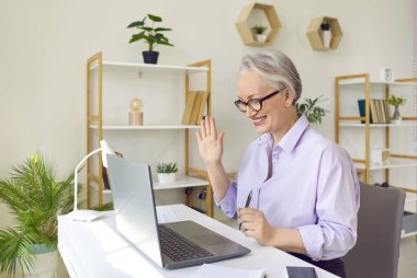 Dost canlısı üst düzey iş kadını dizüstü bilgisayarla çevrimiçi konferansa katılanları selamlıyor. Ofisteki kadının iş görüşmeleri ve online pazarlıklar sırasında web kamerasına el sallamasının yan görüntüsü.