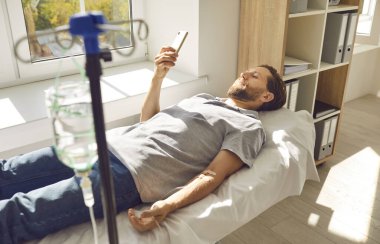 Erkek hasta klinikte damar içi vitamin tedavisi görürken cep telefonu kullanıyor. Adam tıbbi kanepede uzanıyor, serum takılıyor ve modern cep telefonuna mesaj atıyor.