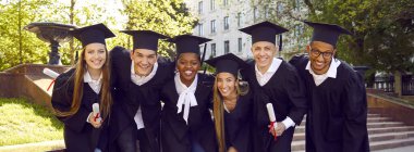 Yeşil kampüs bahçesinde bir grup mutlu üniversite mezunu. Mezuniyet şapkaları ve cüppeleri içinde birkaç neşeli çoklu etnik öğrenci bir arada duruyor, sarılıyor, kameraya bakıyor ve gülümsüyor.