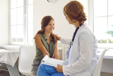 Dost canlısı doktor çocuk hasta destekliyor. Çocuk doktoru olarak çalışan arkadaş canlısı bir kadın sandalyede oturuyor, mutlu, gülümseyen bir liseli kızla konuşuyor, onu rahatlatıyor ve omzuna dokunuyor.