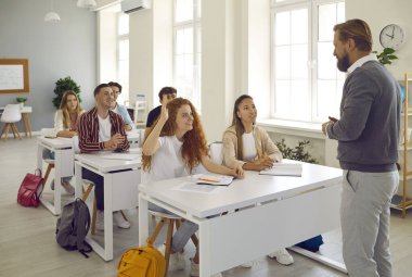 Bir grup lise öğrencisinin dersi var. Sınıftaki beyaz masalarda oturan mutlu genç oğlanlar ve kızlar öğretmenin onlara sorduğu soruyu cevaplamak için ellerini kaldırıyorlar.