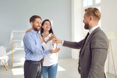 Mutlu genç evli çift gelecekteki yeni bir dairenin anahtarını kabul ediyor. Gülümseyen emlakçı, mutlu müşterileri ev aldıkları için tebrik ediyor. Satılık gayrimenkul kredisi.