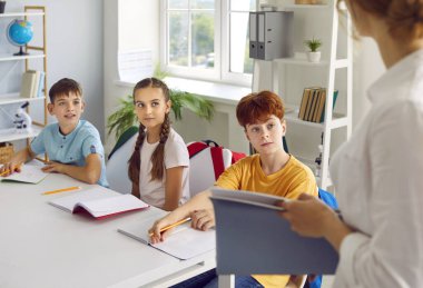 Şirin gülümseyen okul çağındaki çocuklar ferah rahat sınıftaki öğretmenlerini dikkatle dinliyorlar. Masalarında kalem ve okul defterleriyle oturuyorlar. Arka taraftaki öğretmenlerden..