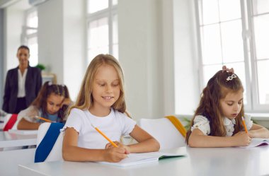 Bir grup ilkokul öğrencisi sınıfta yazıyor. Sınıftaki beyaz masalarda oturan mutlu küçük öğrenci kızlar ellerinde kalemlerle defterlerine yazı yazıyorlar. Okul konseptine dönüş