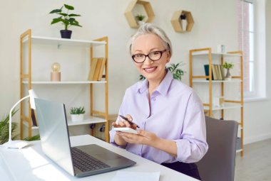 Ofiste çalışan zarif bir iş kadını. Gömlekli, gözlüklü, kısa beyaz saçlı, dizüstü bilgisayarlı, elinde kalem ve cep telefonu olan, kameraya bakıp gülümseyen olgun bir kadın.