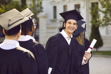 Mutlu genç adam mezuniyetinde eğleniyor. Neşeli, neşeli bir üniversite öğrencisinin açık hava portresi ya da diğer öğrenciler arasında duran kep ve cübbeli üniversite mezununun diploması ve gülümsemesi.