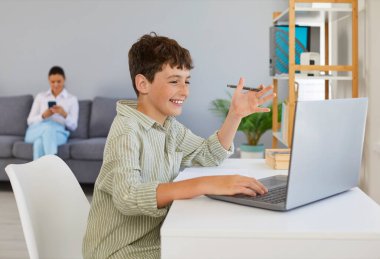 Evde ders çalışan neşeli çocuk video kullanarak öğretmenle ya da öğretmenle konuşuyor. Gülümseyen reşit çocuk dizüstü bilgisayarın önünde oturuyor. Annesinin arka planda oturduğu koltukta cep telefonuyla oturuyor..