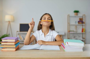 Komik öğrencinin portresi. Masasında kitap ve defterlerle oturan mutlu küçük bir kız çocuğu. Burun altında kalem tutuyor, komik bir yüz ifadesi takınıyor ve parmağıyla işaret ediyor. Okul konseptine dönelim.