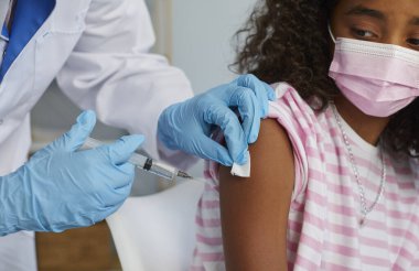 Hemşire kıvırcık saçlı küçük kızın omzuna iğne yapıyor.