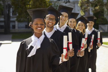 Mezuniyet elbisesi giymiş çok kültürlü insanların gülüşü kampüste kameraya bakıyor. Birbirlerinin arkasında sıraya girerler. Üniversite mezunu, üniversite konsepti.