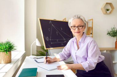 Sınıfta bir kadın matematik öğretmeninin portresi. Gömlekli ve zarif gözlüklü orta yaşlı güzel bir kadın çalışma masasında oturmuş kağıda not alırken kameraya bakıyor.
