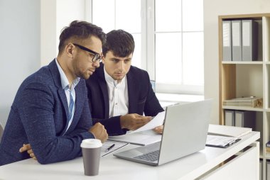 Ofislerinde çalışırken modern bir dizüstü bilgisayar kullanan iki iş adamı takımı. Takım elbiseli iki ciddi genç ofis masasında oturup birlikte bilgisayarlarına bakıyorlar.