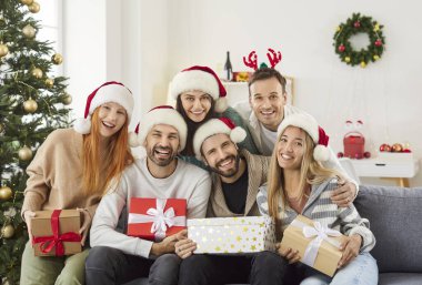 Mutlu genç bir insanın portresi. Evdeki kanepede oturmuş Noel ağacı süslenmiş, hediye kutularını elinde tutuyor ve yeni yılı kutlayan kameraya bakıyor.