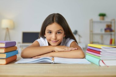 Düşünceli, sevimli, gülümseyen liseli kız kitapların arasında oturmuş kameraya bakıyor ve ellerini defterin üzerine koyuyor. Eğitim, ödev, öğrenim, evde öğrenim, evde eğitim konsepti.