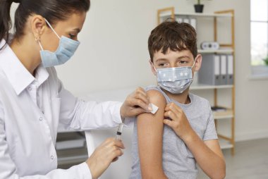 Doktor kadın, klinikte omuz omuza genç bir çocuğa grip aşısı yapıyor. Sağlık, aşı, bağışıklık konsepti. Hemşire ve hasta koruyucu tıbbi maskeler takıyor..