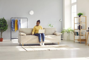 Genç Afrikalı Amerikalı bir kadın evdeki bej renkli koltukta rahat bir şekilde otururken bilgisayarını uzaktan kontrol etmek için kullanıyor. İçi aydınlık, raflar ve elbise raflarıyla dolu bir oturma odasında.