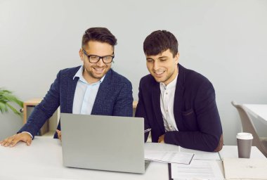 İki arkadaş canlısı erkek arkadaş ortak başlangıç projesinde birlikte çalışıyorlar. Gömlek ve ceket giymiş gülümseyen gençler dizüstü bilgisayarlarıyla masada oturmuş iş planlarını tartışıyorlar..