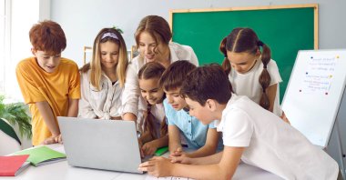 İlkokuldaki çocuklar ve öğretmenleri sınıfta dizüstü bilgisayarda birlikte çalışıyorlar. Masanın yanında duran bilgisayar okur yazarlığının temellerini çalışırlar. Teknoloji kavramını kullanan eğitim.