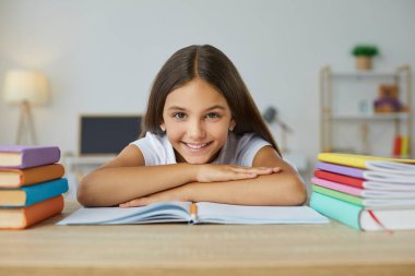 Komik, neşeli ve tatlı liseli kız ders kitapları ve defterlerin arasında oturuyor. Gülümseyen beyaz çocuk portresi, uzanmış, ellerine yaslanmış, kameraya bakıyor..