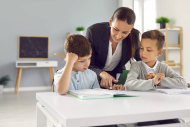 Okul eğitimi. Değer veren bayan öğretmen küçük öğrencilerin okumayı öğrenmesine yardımcı olur ve eğitici materyalleri açıklar. Kadın, elinde kitaplarla masada oturan iki okul çocuğuna doğru eğiliyor..