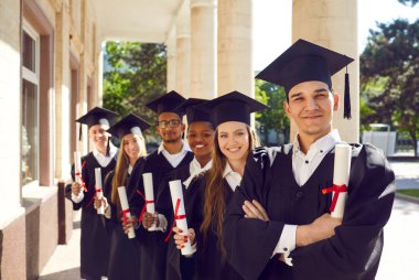 Kampüs bahçesindeki mutlu mezunların toplu portresi. Mezuniyetten sonra elinde kağıt parşömenlerle üniversite binasının yanında durmuş kameraya bakıp gülümseyen altı uluslararası öğrenci.