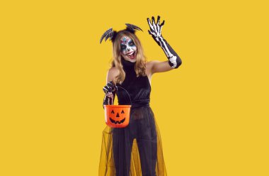 Şeker mi şaka mı? Yaratıcı kostümlü kız Cadılar Bayramı eğlencesinde korkutur, saldırır ve tırmalar. Makyajlı ve Cadılar Bayramı kostümlü genç kız elinde turuncu arka planda şeker kovası tutuyor..