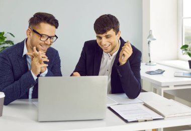 Ofiste çalışırken dizüstü bilgisayar kullanan başarılı iş adamları. Takım elbiseli iki genç ofis masasında oturmuş modern bilgisayar ekranına bakıyor ve gülümsüyor.