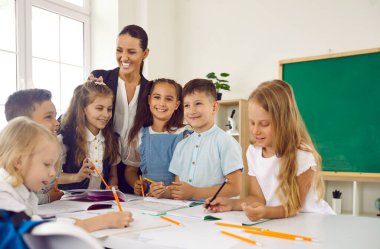 Küçük mutlu çocuklar, ilkokulda okuyan kalemlerle resim çizer kadın öğretmenlerle masanın etrafında durur ve tahtayla birlikte sınıfta iyi bir eğitim alırlar. bilgi, öğrenme