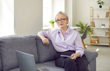 Evdeki kanepede oturan zarif, orta yaşlı bir iş kadınının portresi. Gözlüklü, kısa beyaz saçlı, mutlu, kendinden emin, olgun bir kadın oturma odasında dizüstü bilgisayarıyla kanepede oturuyor.