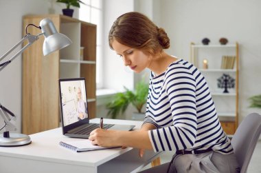 Genç bir kadın dizüstü bilgisayarla öğreniyor, masasında oturmuş notlarını dinliyor ve dinliyor. Mesafe, online eğitim konsepti. Öğrenci üniversitede ders çalışıyor..