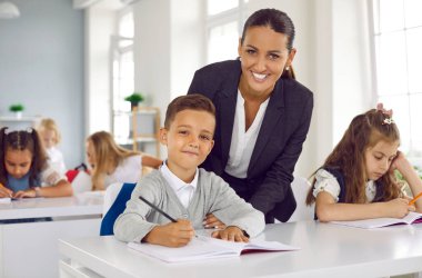 Sınıftaki mutlu, gülümseyen öğretmen ve çocuğun portresi. Genç bir kadın masa başındaki küçük öğrenciye eğiliyor ve ödevini yazmasına yardım ediyor. Okula dönmek, eğitim kavramı.
