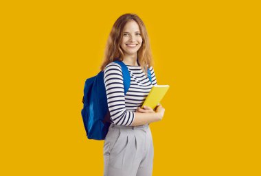 Sırt çantası ve turuncu arka planda ders kitabı olan neşeli kız öğrencinin portresi. Omuzlarında mavi bir sırt çantası olan güzel genç kız kameraya bakıyor ve gülümsüyor. Okul konseptine dönelim..