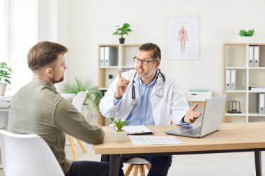 Hastanedeki kendine güvenen erkek doktor dizüstü bilgisayar ekranında muayene sonuçlarını gösteriyor ve klinikte oturan genç bir hastaya danışmanlık yapıyor. Tıp konsepti.