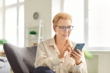 Olumlu yaşlı bir kadın boş zamanlarını cep telefonuyla geçiriyor. İnternette online eğlencenin tadını çıkarmak, sıcak koltukta dinlenmek ve dijital dünyayla iç içe olmak..